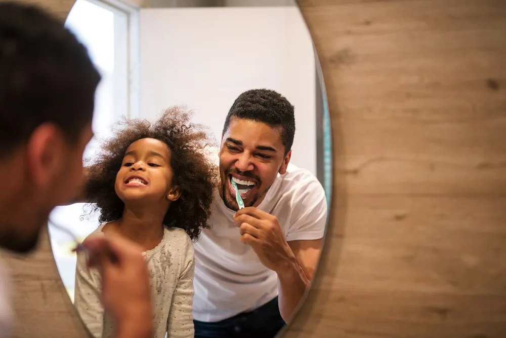 Father and Daughter Brushing Teeth in Front of Mirror – Kirkland Dental Joyful father and young daughter brushing their teeth together while smiling into a mirror – Kirkland Dental