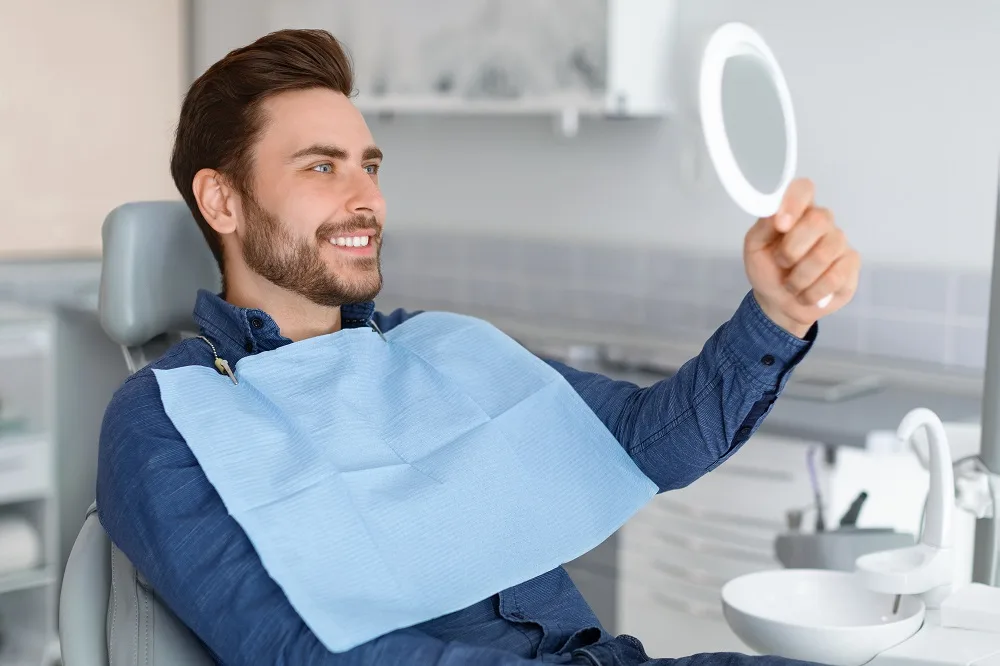 Young man sitting in a dental chair, smiling and holding up a mirror to admire his dental crown results – Dental Crown Kirkland