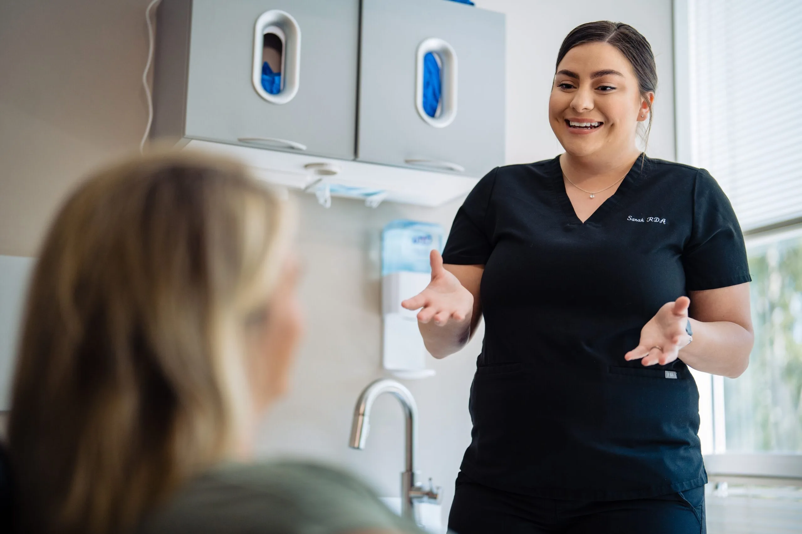 Friendly Dental Assistant Discussing Treatment Plan – Kirkland Dental Smiling dental assistant in black scrubs explaining a procedure to a seated patient inside a bright, modern dental clinic – Kirkland Dental