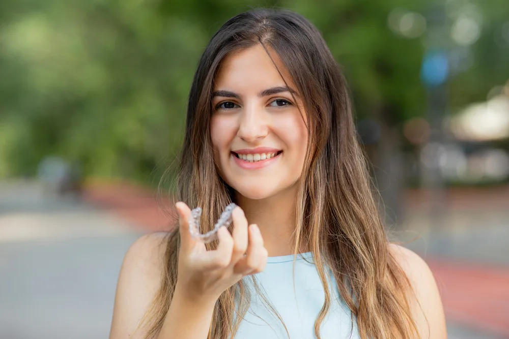 Smiling Woman Displaying Invisalign Tray Outdoors – Kirkland Invisalign Dentist Confident young woman in light blue top holding clear Invisalign aligner with trees in the background – Kirkland Invisalign Dentist