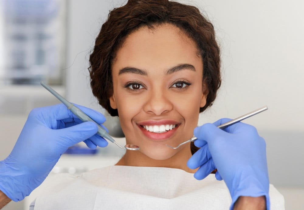 Portrait of smiling african lady and cropped dentist hands in blue gloves with tools in