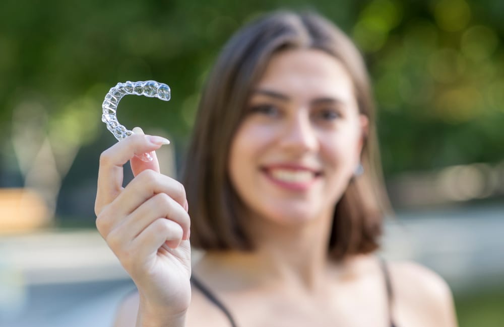 Smiling Woman Holding Clear Aligners – Clear Aligners Kirkland Young woman smiling outdoors while holding a set of clear aligners, promoting discreet orthodontic treatment – Clear Aligners Kirkland