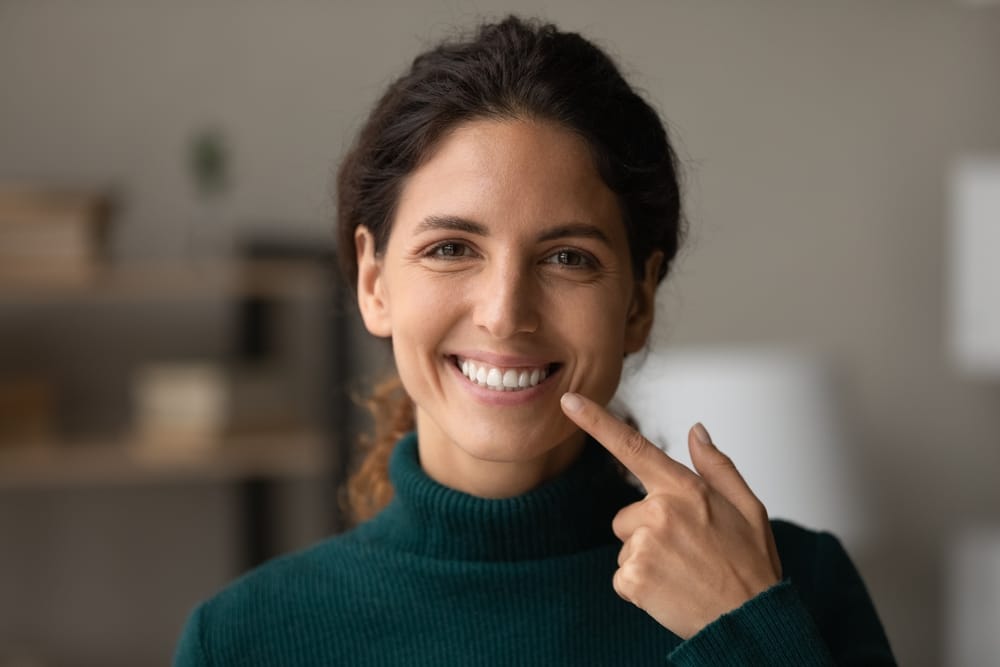 Confident Woman Showing Off Her Smile – Dental Crown Kirkland Smiling woman in green sweater pointing at her teeth, highlighting dental restoration results – Dental Crown Kirkland