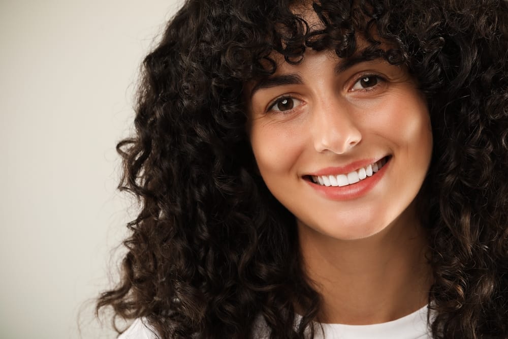 Woman with smooth skin and natural glow – Dermal Fillers Kirkland Smiling woman with radiant, youthful skin and curly hair showing results from - Dermal Fillers Kirkland